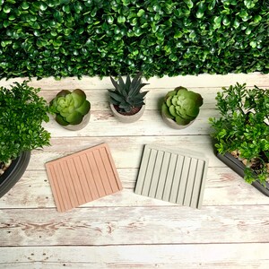 Puede incluir: Dos posavasos rectangulares de hormig&oacute;n con un dise&ntilde;o acanalado. Los posavasos son de color rosa claro y gris claro. Los posavasos est&aacute;n sobre una superficie de madera blanqueada con tres peque&ntilde;as suculentas en macetas.