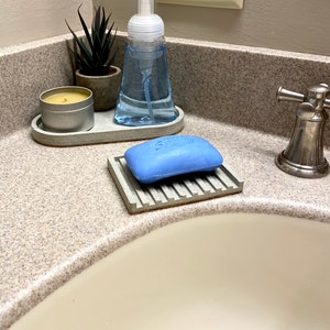 Puede incluir: Un tocador de ba&ntilde;o con un lavabo de porcelana blanca. Un jabonera de hormig&oacute;n gris con una pastilla de jab&oacute;n azul. Una bandeja de hormig&oacute;n gris con una botella de jab&oacute;n transparente y una peque&ntilde;a planta suculenta en maceta. Un portavelas de hormig&oacute;n gris con una vela blanca.
