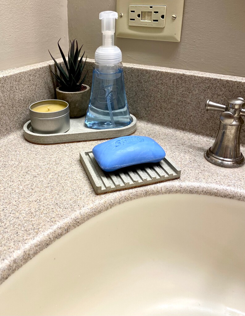 Puede incluir: Un tocador de ba&ntilde;o con un lavabo de porcelana blanca. Un jabonera de hormig&oacute;n gris con una pastilla de jab&oacute;n azul. Una bandeja de hormig&oacute;n gris con una botella de jab&oacute;n transparente y una peque&ntilde;a planta suculenta en maceta. Un portavelas de hormig&oacute;n gris con una vela blanca.