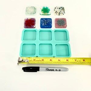 Op de afbeelding: Een turquoise siliconen mal met acht vierkante holtes, ontworpen voor het maken van harsambachten. De mal wordt getoond met een meetlint en een zwarte Sharpie pen.