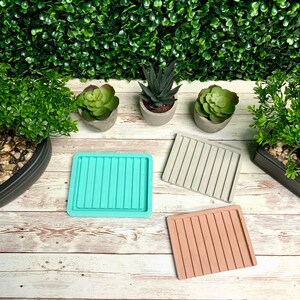Puede incluir: Tres posavasos cuadrados de hormig&oacute;n con un dise&ntilde;o acanalado. Los posavasos son de color turquesa, gris y marr&oacute;n claro. Los posavasos est&aacute;n sobre una superficie de madera blanca.