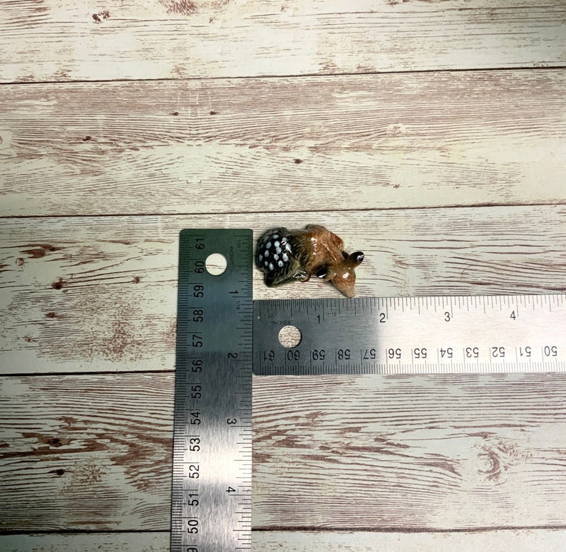 May include: A small ceramic figurine of a brown and white spotted animal, possibly a deer, lying down. The figurine is about 2 inches long and is shown next to a ruler for scale.