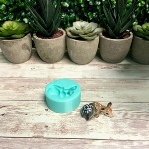 May include: A turquoise silicone mold shaped like a fawn, with a small ceramic fawn figurine next to it. The mold and figurine are on a whitewashed wooden surface.