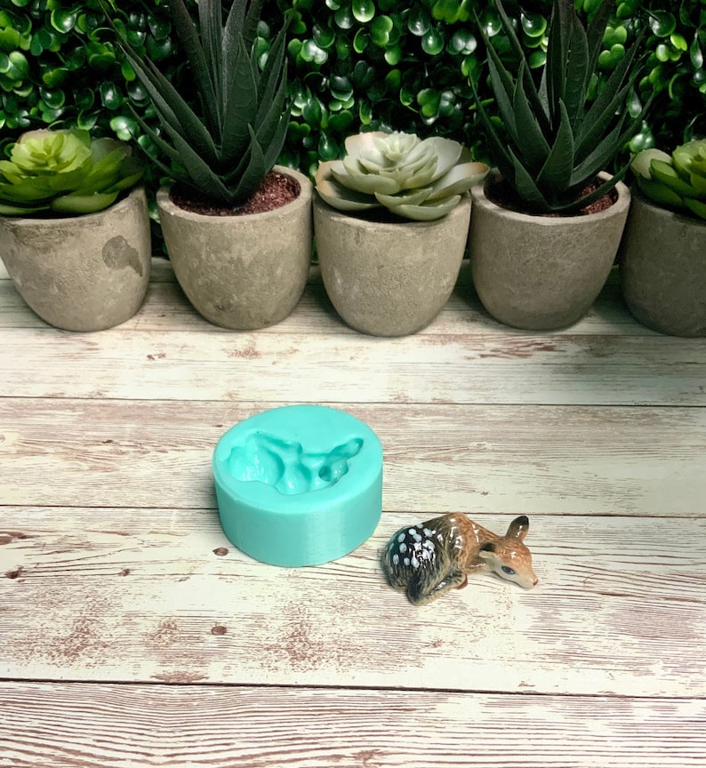 May include: A turquoise silicone mold shaped like a fawn, with a small ceramic fawn figurine next to it. The mold and figurine are on a whitewashed wooden surface.