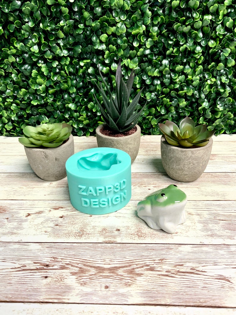Może przedstawiać: Turkusowa silikonowa forma z napisem "ZAPP3D DESIGN" z przodu. Forma ma kształt żaby. Mała ceramiczna figurka żaby stoi na drewnianej powierzchni obok formy.