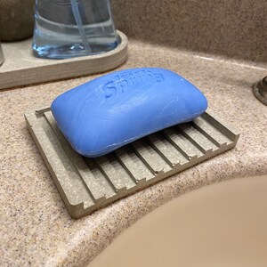 Puede incluir: Una pastilla de jab&oacute;n azul con la palabra "Spring" en relieve, descansando sobre un jabonera de hormig&oacute;n gris con un dise&ntilde;o acanalado.