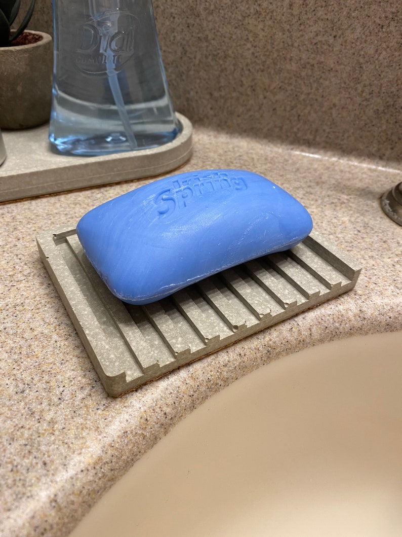 Puede incluir: Una pastilla de jab&oacute;n azul con la palabra "Spring" en relieve, descansando sobre un jabonera de hormig&oacute;n gris con un dise&ntilde;o acanalado.