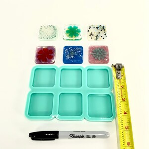 Op de afbeelding: Een set van negen vierkante vormen van helder hars, waarvan er zes zijn gevuld met gekleurd hars en glitter. De harsvierkanten zijn ongeveer 2,5 cm bij 2,5 cm. Een liniaal en een zwarte Sharpie-marker zijn ook op de afbeelding te zien.