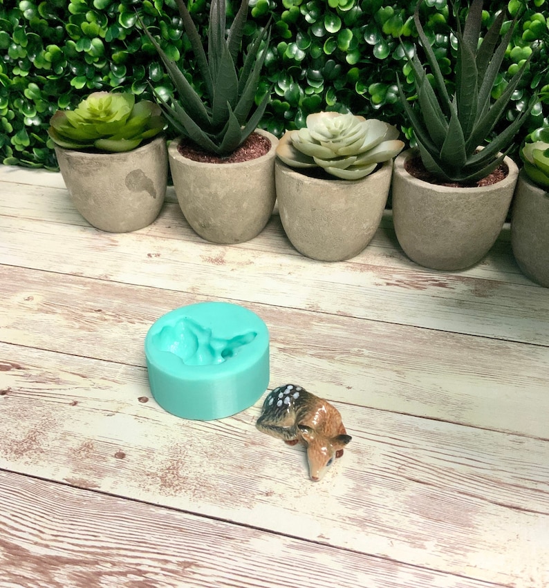 May include: A turquoise silicone mold shaped like a deer, with a small ceramic deer figurine next to it. The mold is on a light wood surface.