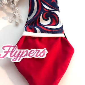 Op de afbeelding: Een rood en blauw patroonaccessoire met het woord "Flypers" in roze script. Het bovenste gedeelte heeft een wervelend ontwerp in rood, wit en blauw, met blauwe bandjes. De onderkant is effen rood.