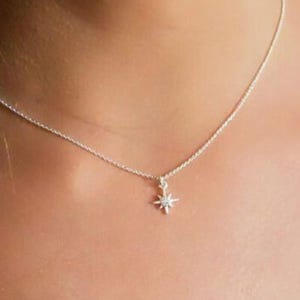 May include: A delicate silver necklace featuring a small starburst pendant with a central gemstone. The pendant hangs from a fine chain, creating a minimalist and elegant look. The necklace is designed to be worn close to the neck.
