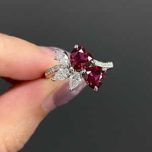 May include: A silver ring featuring a cluster of white, pear-shaped stones and two heart-shaped, deep red gemstones. The ring has a delicate, elegant design, perfect for a special occasion.