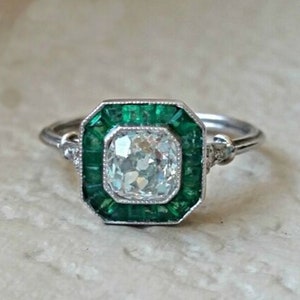May include: A silver ring with a large, square-cut diamond surrounded by a halo of emerald green stones.