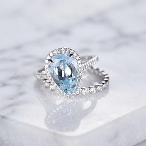Può includere: Un anello color argento con una grande pietra centrale azzurra a forma di goccia, circondata da un alone di piccole pietre rotonde trasparenti. La fascia è decorata con una fila di pietre trasparenti abbinate per un aspetto scintillante.