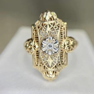 May include: A gold filigree ring with a central floral design and a small, clear gemstone. The ring has an ornate, openwork design with decorative elements on the sides. The ring is set against a neutral background.