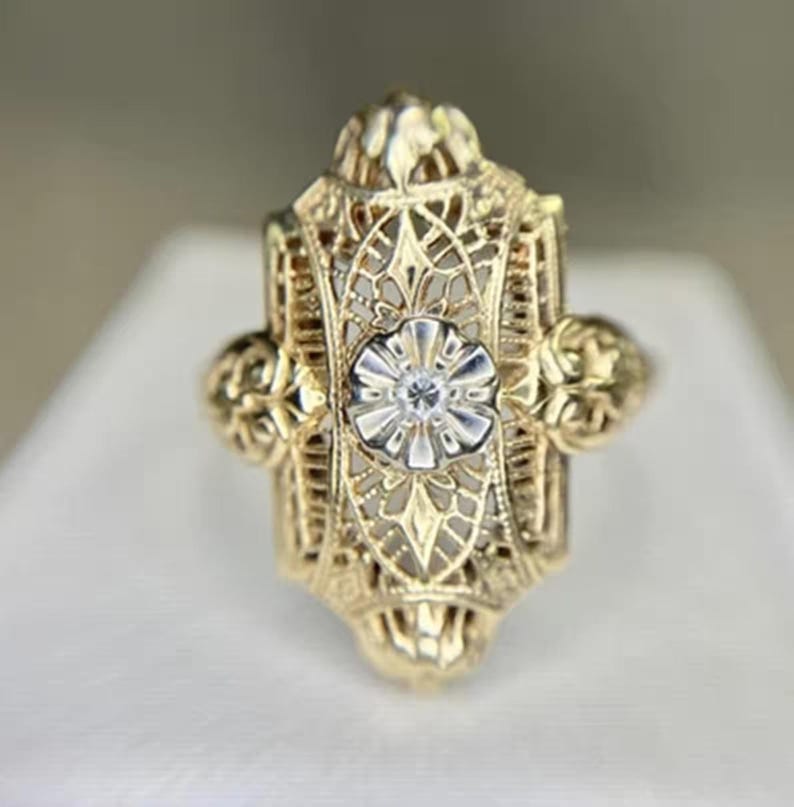 May include: A gold filigree ring with a central floral design and a small, clear gemstone. The ring has an ornate, openwork design with decorative elements on the sides. The ring is set against a neutral background.
