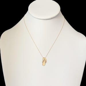 May include: A gold-colored necklace featuring a small flip-flop pendant. The pendant has a textured surface and is adorned with small, clear gemstones. The necklace is displayed on a white mannequin against a black background.
