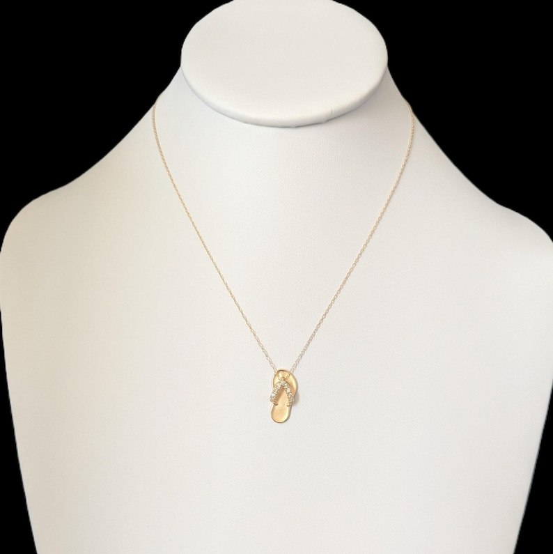 May include: A gold-colored necklace featuring a small flip-flop pendant. The pendant has a textured surface and is adorned with small, clear gemstones. The necklace is displayed on a white mannequin against a black background.