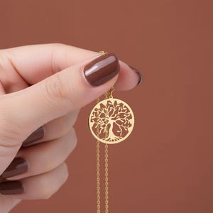 May include: A gold-colored pendant necklace featuring a circular tree of life design. The necklace is held against a brown background, showcasing the intricate details of the pendant and the delicate chain.