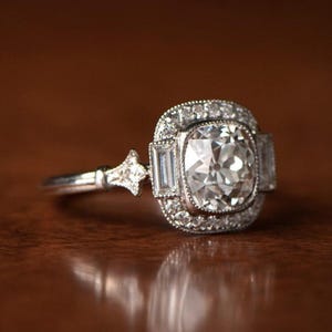 May include: A silver engagement ring with a large, round, clear diamond surrounded by smaller diamonds. The ring has a square-shaped setting with a halo of smaller diamonds.