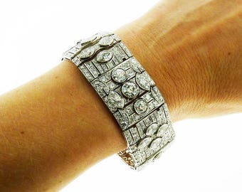 May include: A wide, ornate bracelet with a repeating pattern of round and oval-shaped clear gemstones set in a silver-colored metal. The bracelet is worn on a wrist, and the gemstones sparkle against the skin.