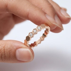 May include: A rose gold ring with a honeycomb design. The ring features a series of hexagonal shapes, with three of the hexagons containing small, sparkling diamonds. The ring is held between two fingers against a white background.