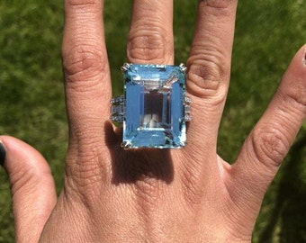 May include: A large, rectangular aquamarine gemstone ring set in white gold with a diamond band. The ring is on a person's finger.