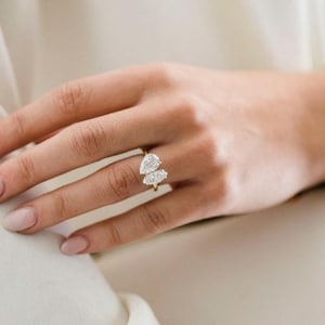 K&ouml;nnte beinhalten: Ein goldener Ring mit zwei birnenf&ouml;rmigen Diamanten, die auf einem Band sitzen.