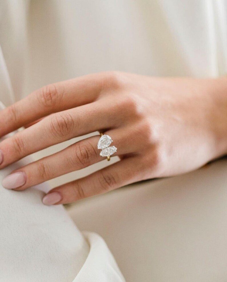 K&ouml;nnte beinhalten: Ein goldener Ring mit zwei birnenf&ouml;rmigen Diamanten, die auf einem Band sitzen.
