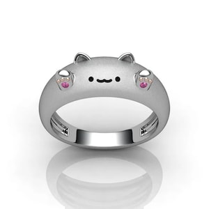 May include: A silver ring with a cat face design. The cat has pink paw prints on its cheeks and a small pink gem in each paw print.