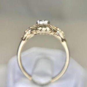 May include: A gold-colored ring with a small, clear gemstone at the center. The ring features intricate floral detailing around the gemstone and along the band. The ring is set against a soft, neutral background.