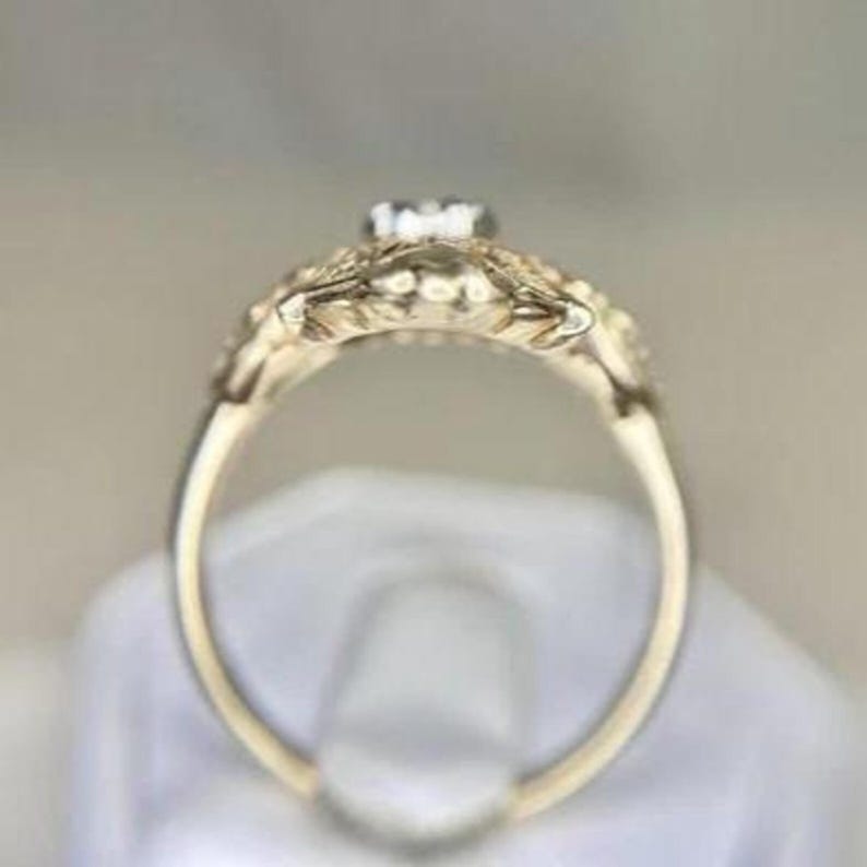 May include: A gold-colored ring with a small, clear gemstone at the center. The ring features intricate floral detailing around the gemstone and along the band. The ring is set against a soft, neutral background.