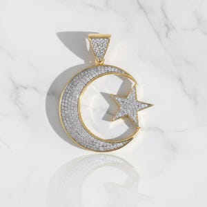 Op de afbeelding: Een goudkleurige hanger met een maan en een ster, beide bezet met sprankelende witte stenen. De hanger heeft een driehoekige beugel voor een ketting. Het ontwerp is geplaatst tegen een witte marmeren achtergrond.