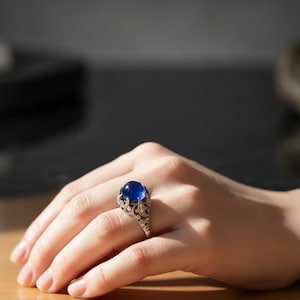 May include: A silver ring with a large, round, deep blue gemstone. The ring has an ornate, filigree design around the gemstone. The ring is on a person's hand, with the background out of focus.