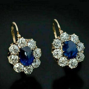 Op de afbeelding: Een paar gouden oorbellen met een blauwe saffier edelsteen omringd door kleinere diamanten. De oorbellen hebben een bloemmotief en hangen aan een haak.