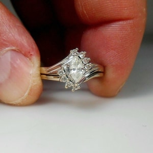 Puede incluir: Un anillo de compromiso de oro blanco con un diamante de talla marquesa y un halo de diamantes más pequeños. El anillo está sobre un fondo blanco.