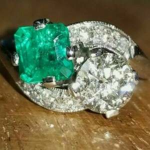 Könnte beinhalten: Ein silberner Ring mit einem großen Smaragd und einem kleineren runden Diamanten. Der Ring ist mit kleineren Diamanten um die Edelsteine besetzt.