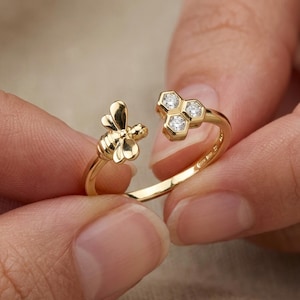 May include: A gold-colored ring featuring a bee and honeycomb design. The bee is on one side, and a honeycomb with three clear gemstones is on the other. The ring is held between two fingers.