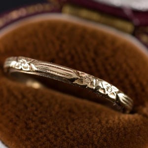 May include: A gold ring with a floral pattern, resting in a brown velvet jewelry box. The ring's surface is detailed with small, embossed flowers and delicate lines. The box has a burgundy border.