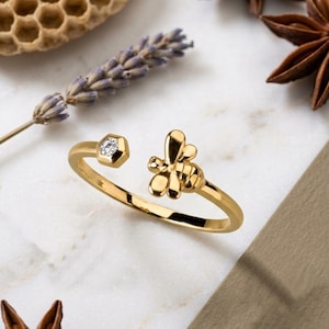 May include: A gold-colored ring with a bee design. The ring features a small diamond in a hexagonal setting and a bee on the opposite end. The ring is set against a backdrop of lavender, star anise, and a honeycomb.