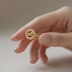 May include: A gold-colored ring with a unique design, featuring a circular setting with two small spheres. The ring is held in a hand, showcasing its intricate details and craftsmanship. The ring's design is reminiscent of a nest.