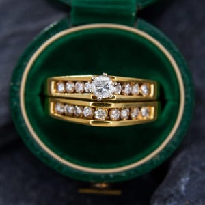 May include: A gold wedding ring set with a large, round, clear diamond center stone and smaller diamonds set along the bands. The rings are displayed in a green velvet ring box.