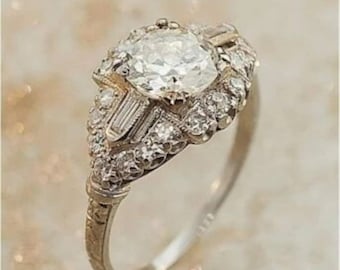 May include: A vintage-style engagement ring with a large, round, clear diamond surrounded by smaller diamonds. The band is made of white gold and has a delicate, intricate design.