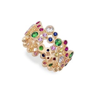 Può includere: Un anello dorato con un gruppo di pietre preziose colorate. L'anello è decorato con gemme rotonde nei toni del verde, rosa, blu, giallo e trasparente. Le gemme sono incastonate in un design strutturato e organico.