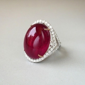 May include: A large, oval-shaped ruby gemstone ring. The ruby is a deep red color and is surrounded by a halo of small, sparkling diamonds. The ring has a silver band with gold accents.