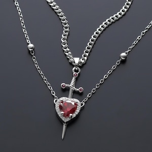 May include: A silver necklace featuring a heart-shaped pendant pierced by a sword. The heart is a deep red gemstone, surrounded by smaller clear stones. The necklace has two chains, one a curb chain and the other a delicate chain with small beads.