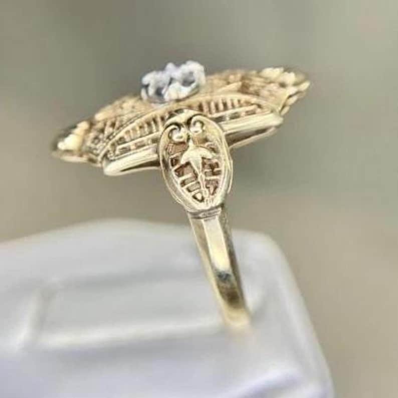 May include: A gold-toned ring with an ornate, filigree design. The ring features a central, clear gemstone and intricate detailing on the band and setting. The ring's design is reminiscent of vintage or antique jewelry.
