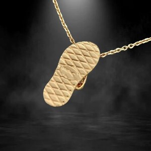May include: A gold-colored pendant necklace featuring a detailed flip-flop charm. The charm has a textured sole design and is suspended from a delicate chain. The pendant is a warm gold tone, and the background is dark.
