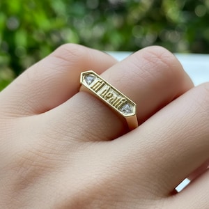 May include: A gold ring with the inscription "til death" in a stylized font. The ring has a rectangular design with a diamond-shaped accent on each end. The ring is worn on a finger.