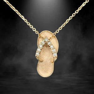 May include: Gold-toned pendant necklace featuring a flip-flop design. The flip-flop has a textured surface and is adorned with small, sparkling gemstones along the straps. The chain is delicate and complements the pendant's design.